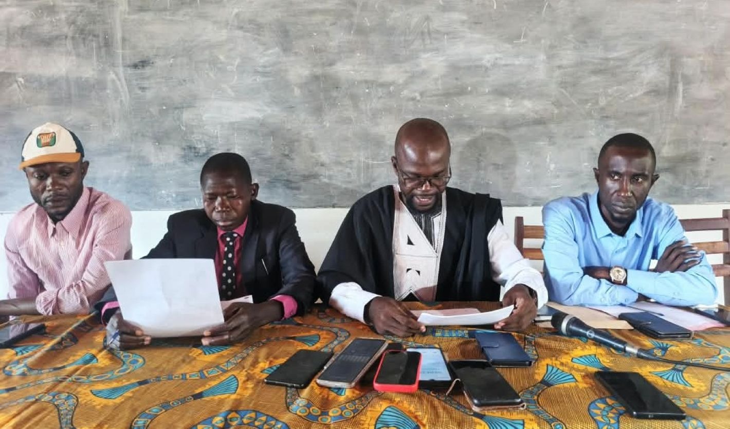 Four men sit behind a table with phones and papers during a press conference in front of a blackboard.