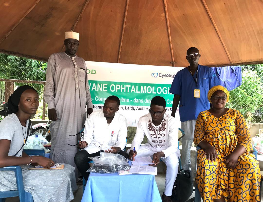 Medical staff and volunteers sit and stand under a canopy in front of an eye-care caravan banner.