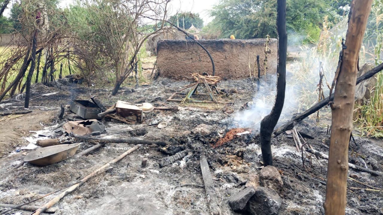 Burned household belongings and smouldering debris in a yard in front of a mud-brick wall.