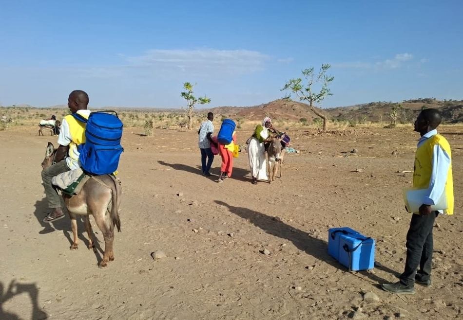 Health workers in yellow vests carry blue cold boxes across dry ground, one on a donkey.
