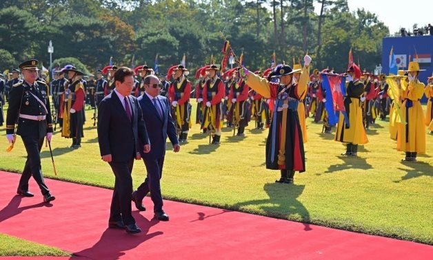 Two men in suits walk along a red carpet past an honour guard in traditional costume.