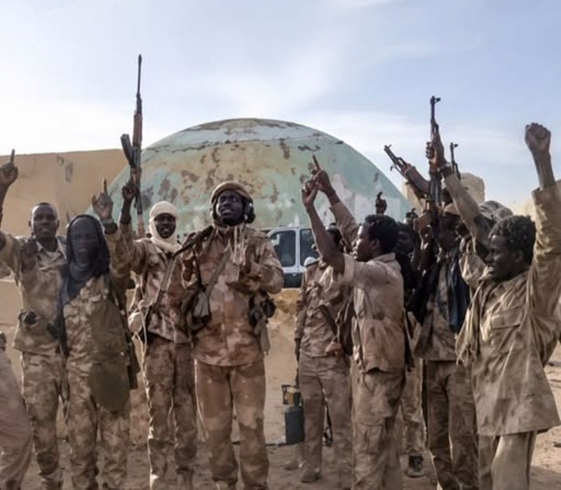 Soldiers in camouflage raise their fists and rifles in front of a domed building.