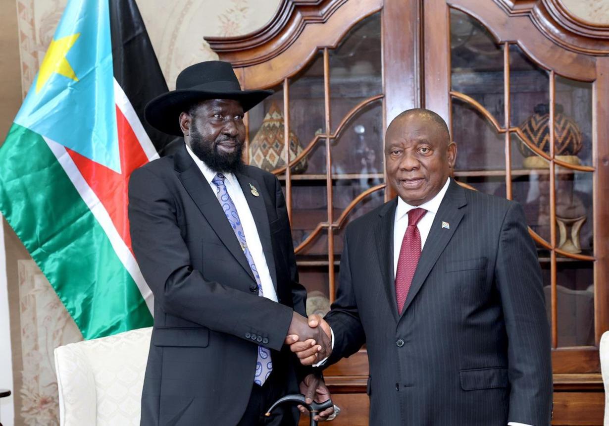 South African President Cyril Ramaphosa and South Sudanese President Salva Kiir shake hands in front of a South Sudanese flag.