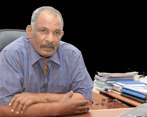 Portrait of an older man in a blue checked shirt, arms crossed on a desk with stacked papers.