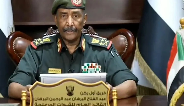 Army commander in uniform and beret seated at a desk with a nameplate and flags behind him.