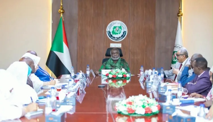 A uniformed official chairs a long meeting table with a Sudanese flag and an emblem on the wall behind him.