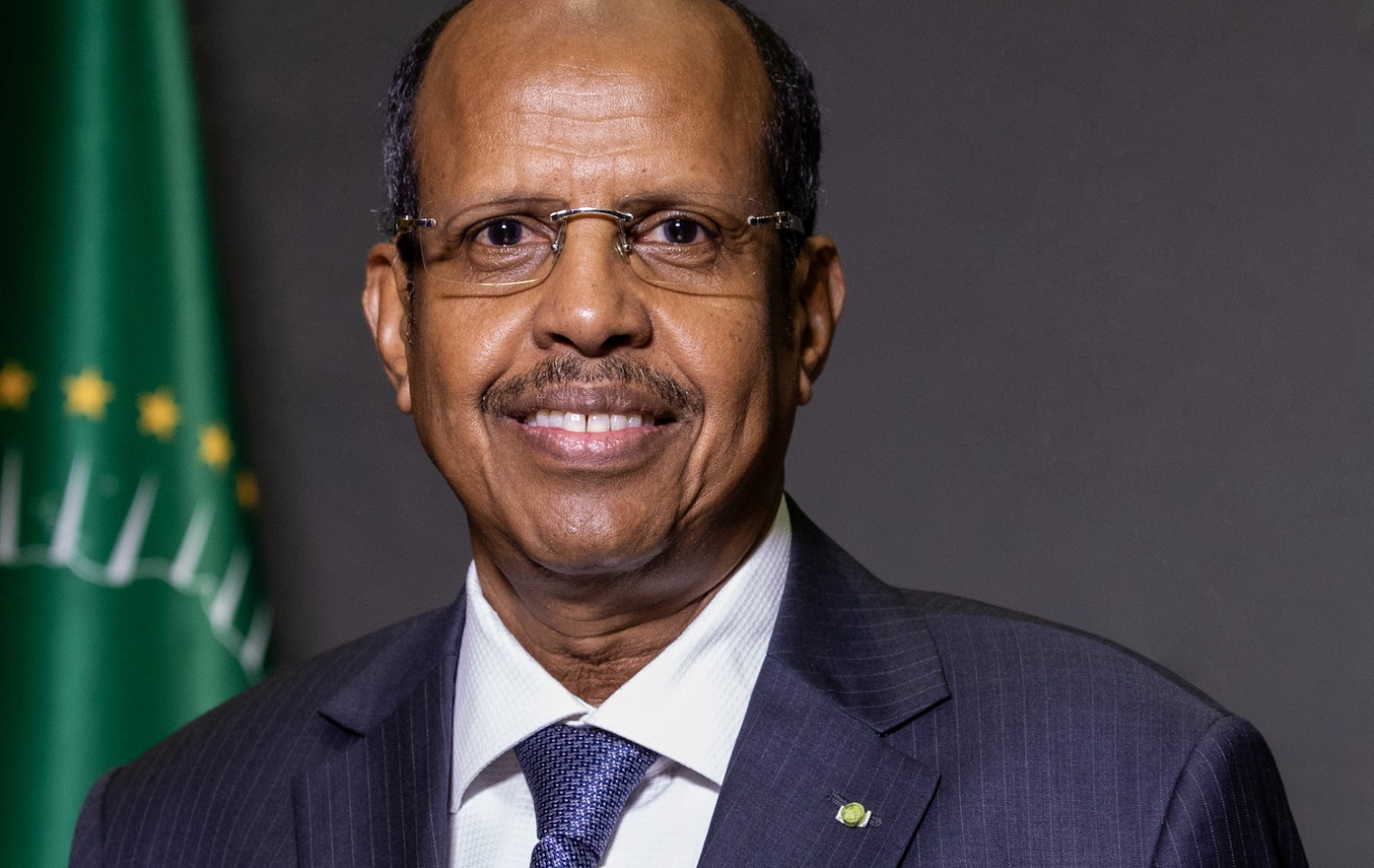 Portrait of a man in glasses and a dark suit smiling in front of an African Union flag.