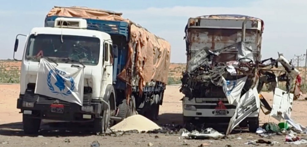 Two trucks on a desert road, one marked with the WFP flag and the other badly wrecked.
