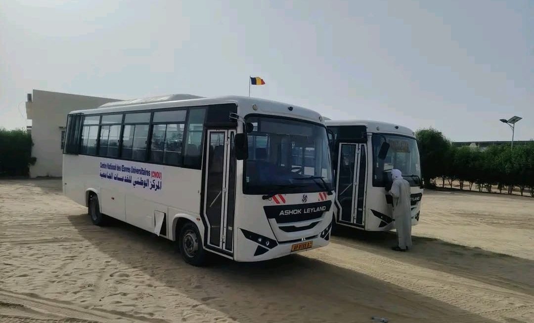 Two white buses parked on sand in front of a low building with a Chadian flag.